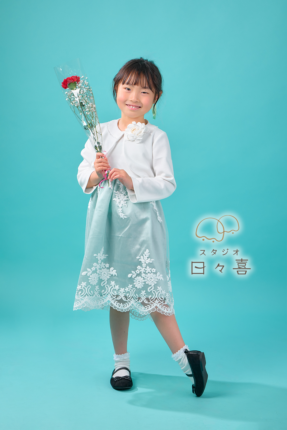 ターコイズブルーの背景の前で、赤いカーネーションの花束を大切そうに抱えて微笑む、白いボレロとミントグリーンのドレスを着た女の子の全身写真。