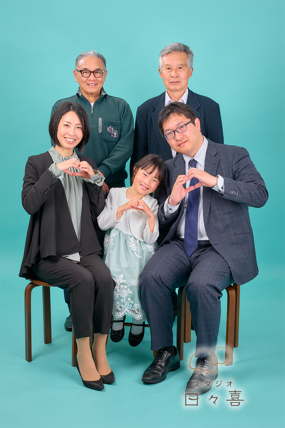 ご両親とお子様の3人が手でハートマークを作って寄り添う、仲睦まじい家族写真。背景には優しく見守る祖父の姿もあり、家族の絆が伝わるカット。